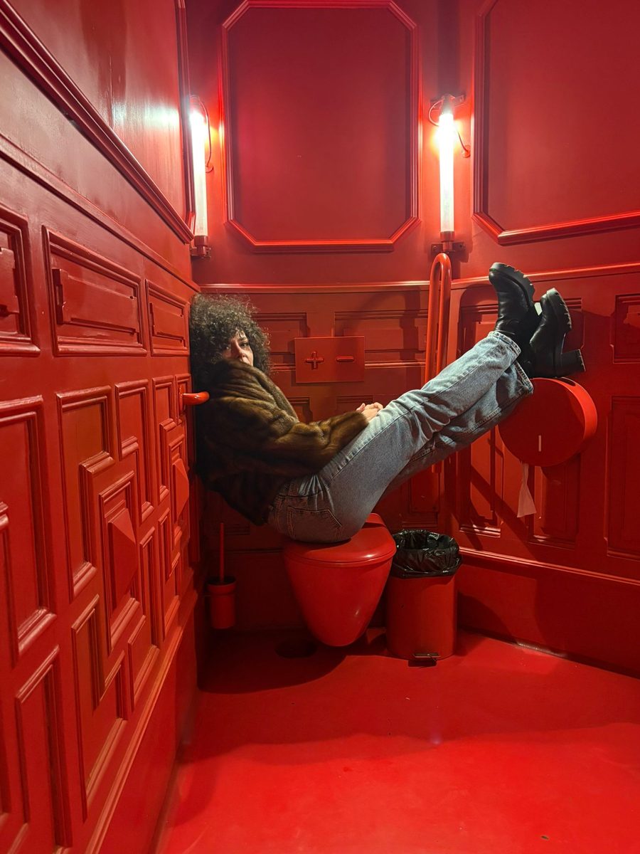 Carlo tumbada sobre el váter de un baño completamente rojo, con abrigo de pelo y las botas apoyadas en la pared